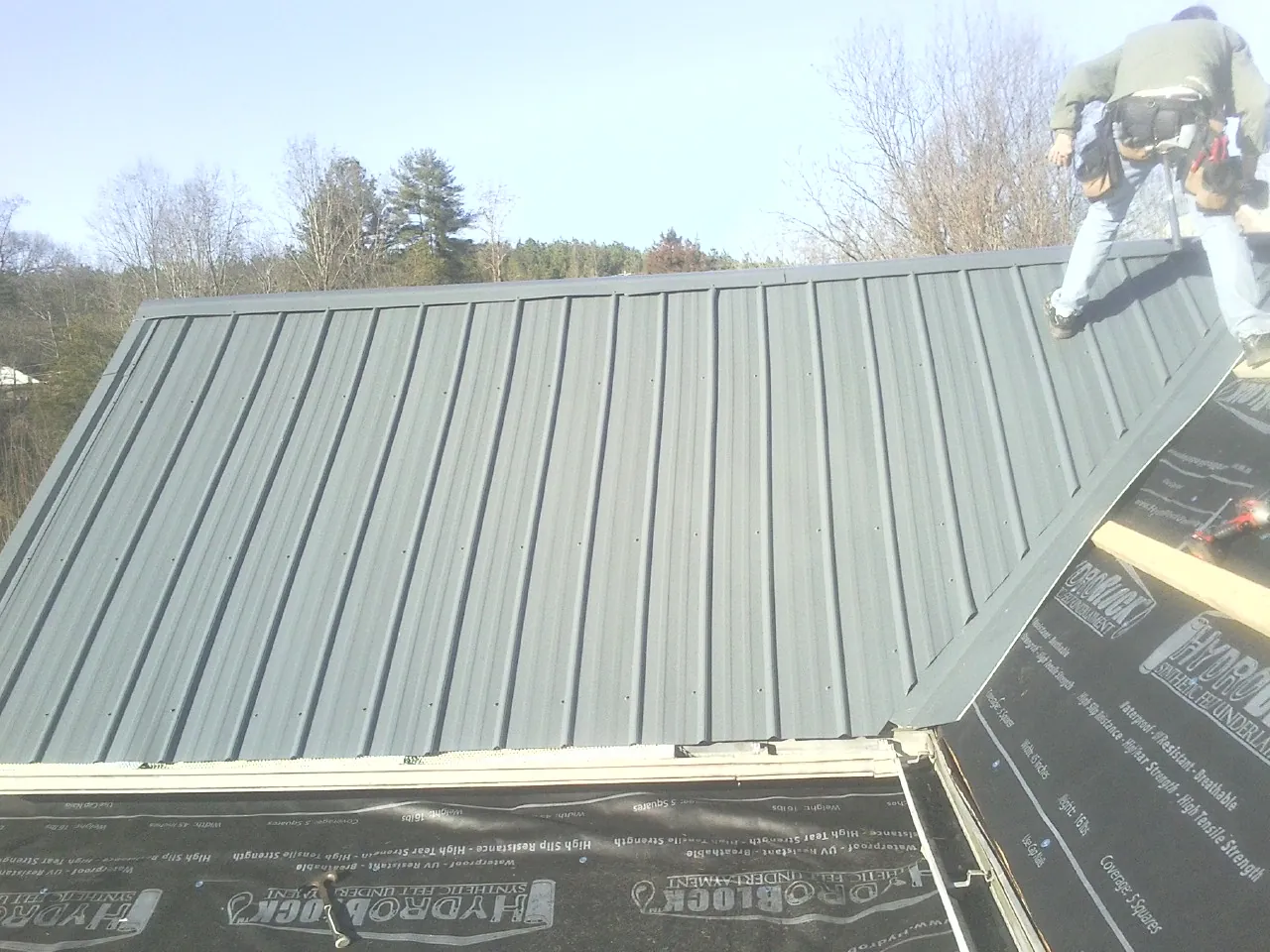 Crew installing dark green standing seam panels over synthetic underlayment in Cranston