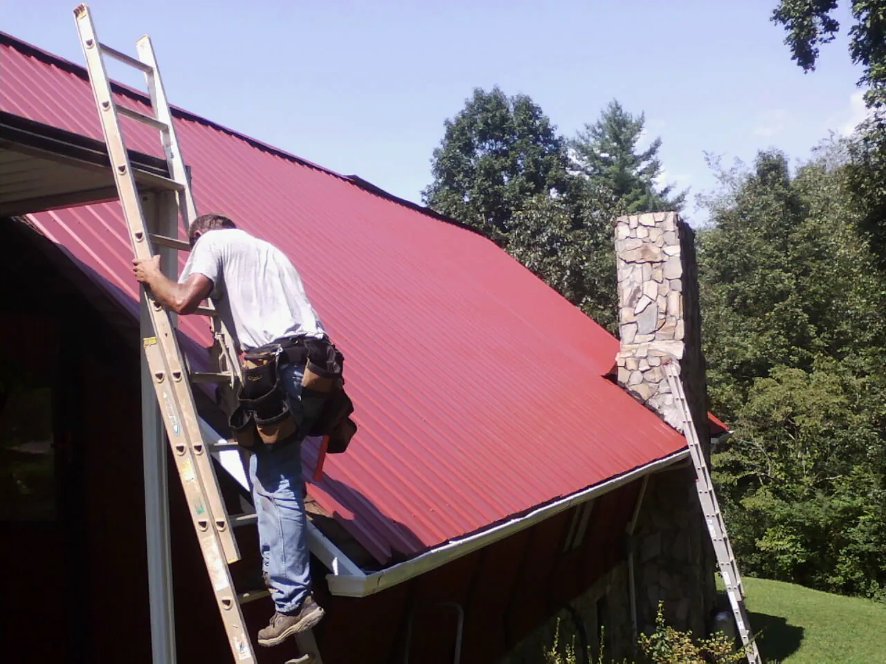 Roofer on a ladder beside a red metal roof and stone chimney — Soffit & Fascia Repair by Roofing Brothers in Cranston